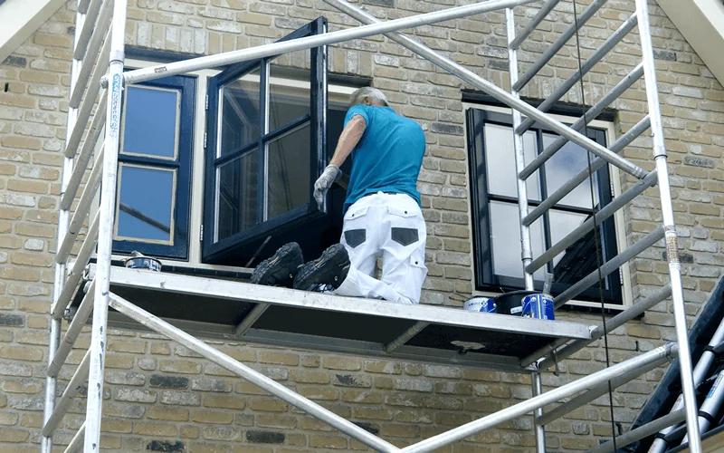 Buitenschilderwerk: schilder behandelt vanaf een steiger de buitenkozijnen van een woning